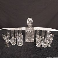 Full set showing the decanter in the center surrounded by 18 assorted shot glasses in varied styles on a black cloth.