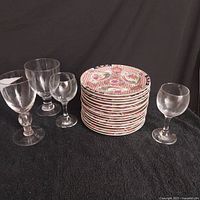 Group of 8 stacked Asian porcelain plates surrounded by 4 clear vintage glasses of different shapes and sizes on a black cloth background.