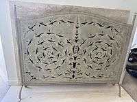Front view of a grey-green metal fireplace cover with intricate cut-out floral and swirl patterns, standing on two legs with horizontal supports.