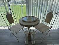 Photo shows a round metal patio table covered with a textured beige cloth, with a shiny round mirror placed on top, flanked by two matching metal chairs with beige floral patterned cushioned seats and backs.