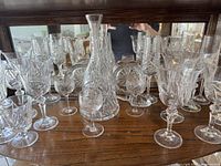 Photo of crystal glassware set displayed in curio cabinet showing variety of wine glasses, sherry glasses, flutes, crystal decanter center, and crystal fruit bowls on shelf below.