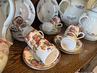 Front view showing 8 demi-tasse cups and saucers stacked on a wooden surface with teapot and other serving pieces in the background.