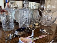 Photo showing crystal decanter, crystal highball glasses, red wine glasses, china fruit bowls and silver cruets, sugar salver with utensils arranged on wooden surface against mirrored back.