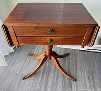 Photo of wooden trestle table showing top surface, two front drawers with knobs, drop leaves folded down, and pedestal legs.