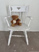 Full frontal view of the antique wooden armchair painted white with wear and paint chipping visible, with a brown teddy bear placed on the seat.