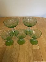 Front and angled view of 5 vintage green glass dessert cups arranged in a pyramid style showing etched leaf pattern and stem bases on a wood table.