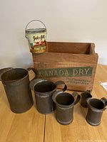 Image showing the Jack & Jill peanut butter can on edge of Canada Dry wooden crate with five dark metal tin measuring cups in front.