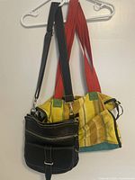 Multicolor Barefoot fabric bag with red shoulder straps alongside smaller black Roots leather crossbody bag hung on hanger.