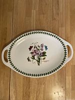 Top view of white oval ceramic plate with integrated side handles, decorated with colorful Sweet Pea flower and butterfly illustration, surrounded by green leaf border.