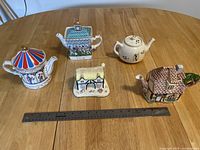 Top-down view of the four decorative teapots and one small cottage figurine arranged on a wooden table with a ruler for scale.