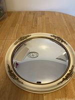 Top view of circular mirror laying flat on a wooden surface. Shows cream-colored wooden frame with decorative floral detailing and reflection of a wall clock.