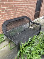 Front view of dark brown wicker outdoor bench with notable wear and overgrown plants around base.
