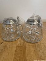 Two clear crystal steins with heavy cut glass design and pewter lids wrapped in clear plastic on wooden surface.