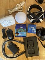 Photo of assortment of electronics including Fujifilm camera, Philips razor, over-ear headphones, Sony neckband speaker, CD-R discs, mouse, phone case, lung expansion device, Klogic electronic reader, Panasonic CD player, and various cables on wooden floor.