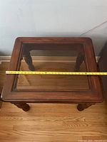 Top view of square wooden side table with glass top and measuring tape showing 24 inch width.