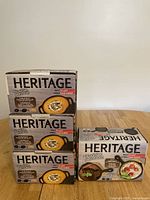 Four unopened boxes of Heritage mini cast iron cookware shown stacked, three mini skillets and one casserole.