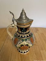 Front angled view showing ornate colorful ceramic body and pewter lid with thumb lever.