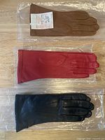 Three pairs of leather gloves in black, red, and brown colors laid flat on a wooden surface, all inside plastic sleeves.