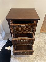 Dark brown wooden drawer chest with three wicker drawers and leather handles, drawers opened showing interior space, minor wear visible on top surface.