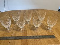 Seven clear crystal goblets arranged in a row on a wooden table with a ruler in front