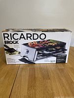 Front view of the boxed raclette grill showing product name, brand, and image of the grill with food on top and raclette pans.