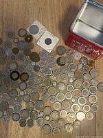 Photo shows assortment of various coins spread on wooden surface next to open tin container.