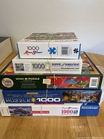 Stack of six boxed jigsaw puzzles from various brands including Ravensburger, Cobble Hill, Bits & Pieces, and Aimee Stewart. Visible puzzle piece counts of 1000 and 500 pieces.