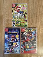 Front cover of the three Nintendo Switch game cases stacked vertically on wood surface showing game titles: New Super Mario Bros. U Deluxe, Mario + Rabbids Sparks of Hope, and Super Mario Odyssey.