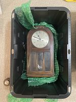 A vintage wooden wall clock with arched top inside a black plastic bin with green bubble wrap.