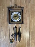Front view of the West German vintage wall clock movement with visible brass dial featuring Roman numerals, black hands, two brass weights and pendulum on chains.