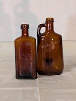 Photo shows two vintage amber glass bottles; one square, one with handle and embossed script.