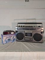 Front view of vintage silver and black cassette player and radio combo with dual large front speakers and a smaller cassette player on its original box placed to its left side