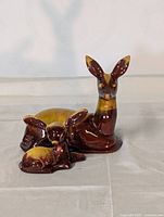 Photo showing ceramic doe figurine seated upright with a smaller fawn lying beside it, both glazed in brown and yellow.
