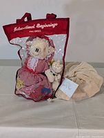 Photo of piggy doll in original packaging with smaller pig doll and mesh bag containing wooden blocks beside it.
