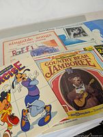 Four children's vintage records on a flat surface including 'Singable Songs' by Raffi with scribbled markings, 'Mousecise' Disney record, 'Country Bear Jamboree', and Anne Murray album.