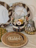 Photo showing fall-themed items: grapevine wreath wrapped in burlap ribbon, decorated basket with faux flowers and pumpkin, silver ribbed pumpkin gourd, and wooden 'thankful' platter.