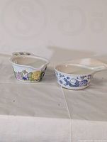 Two vintage Figglo ceramic sauce boats on a white cloth background. One has colorful floral pattern, the other has blue and white pattern with a woman figure.