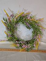 Full view of the circular floral wreath made of artificial flowers and foliage on a white background with ruler at base.