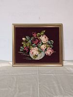 Front view of framed needlepoint floral artwork with roses and green leaves on burgundy background.