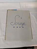 Front cover of a spiral-bound scrapbook titled 'Scrap Book' showing a plain, light gray cover with blue lettering.