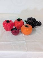 Five colorful glass fruit figurines arranged on a light fabric background, including a grape cluster, apple, orange, bell pepper, and small fruit.