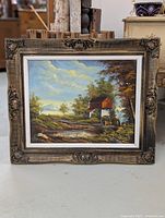 Framed oil painting showing a water mill by pond with trees and sky