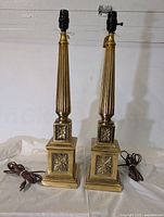 Photo showing full view of two tall brass lamps with fluted bodies and floral decorated square bases with cords and bulb sockets.