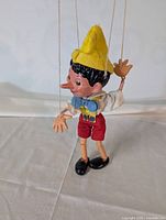 Front view of Pinocchio marionette puppet showing yellow hat, red shorts, and blue bow tie.