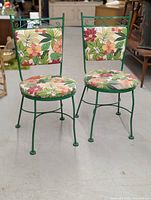 Front view of two green metal chairs with floral-patterned cushions on seat and backrest, showing decorative metalwork and general condition.