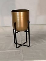 Front angle view of the gold metal planter on black metal stand showing cylindrical pot and square crossbar frame stand on beige floor background.