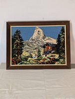 Full view of framed needlepoint artwork showing mountain range, cabin, and trees