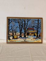 Full view of the framed needlepoint winter scene artwork showing snowy landscape, yellow house, leafless trees, and figure walking