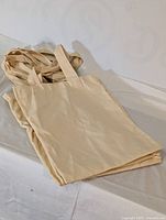 Stack of plain beige tote bags with handles visible, laid flat on a light-colored surface.