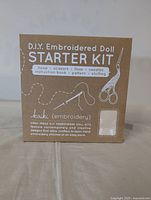 Box of Kurik DIY Embroidered Doll Starter Kit showing product name and included contents such as hoop, scissors, floss, needles, instruction book, pattern and stuffing. Elephant design depicted.
